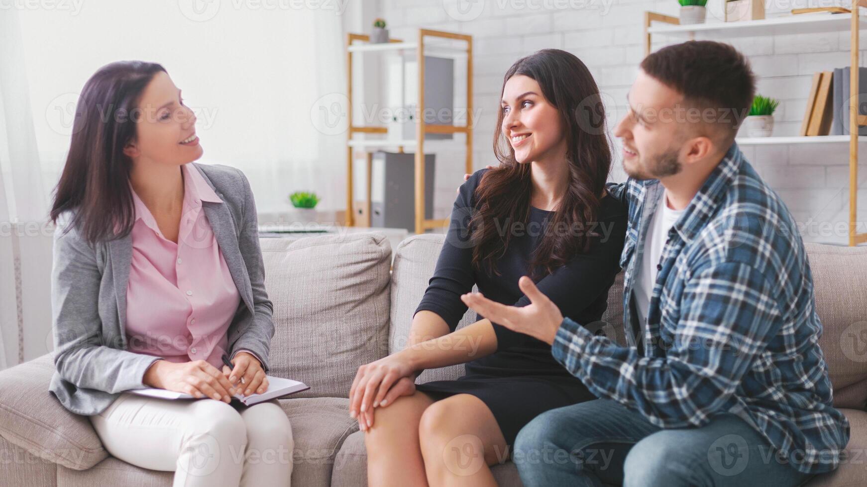 A young couple is sitting on a couch in an office setting, talking with their therapist. The therapist is a woman, and she is listening intently to the couple photo