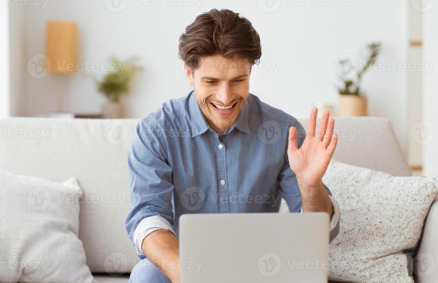 A man sits on a couch, smiling and waving to the laptop screen in front of him. photo