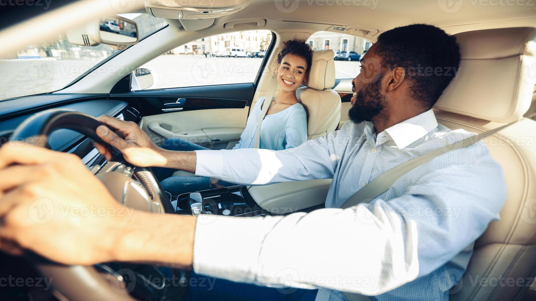 African American man with a beard is driving a car while a woman sits in the passenger seat. He is looking forward, and she is smiling at him. The car is in motion, and they are in a city environment. photo