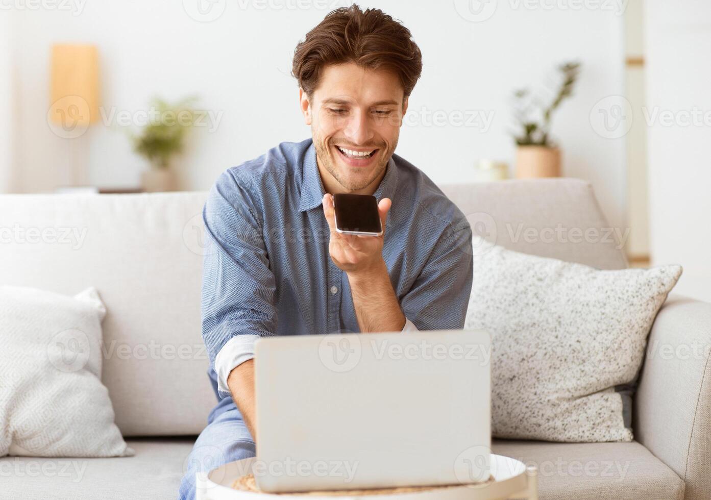 A man is sitting on a couch, smiling while looking at a smartphone in his hand. He appears to be using a voice assistant to complete a task while working on a laptop. photo