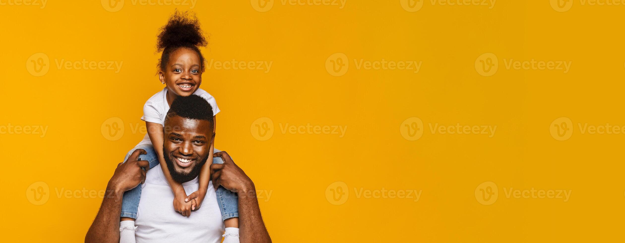 A cheerful black father carries his smiling daughter on his shoulders, both filled with laughter. They are enjoying a moment of playful bonding, highlighted by the vibrant yellow backdrop, copy space photo