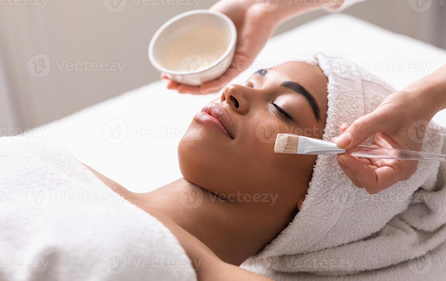 Spa therapist applying face mask on african woman skin, closeup photo