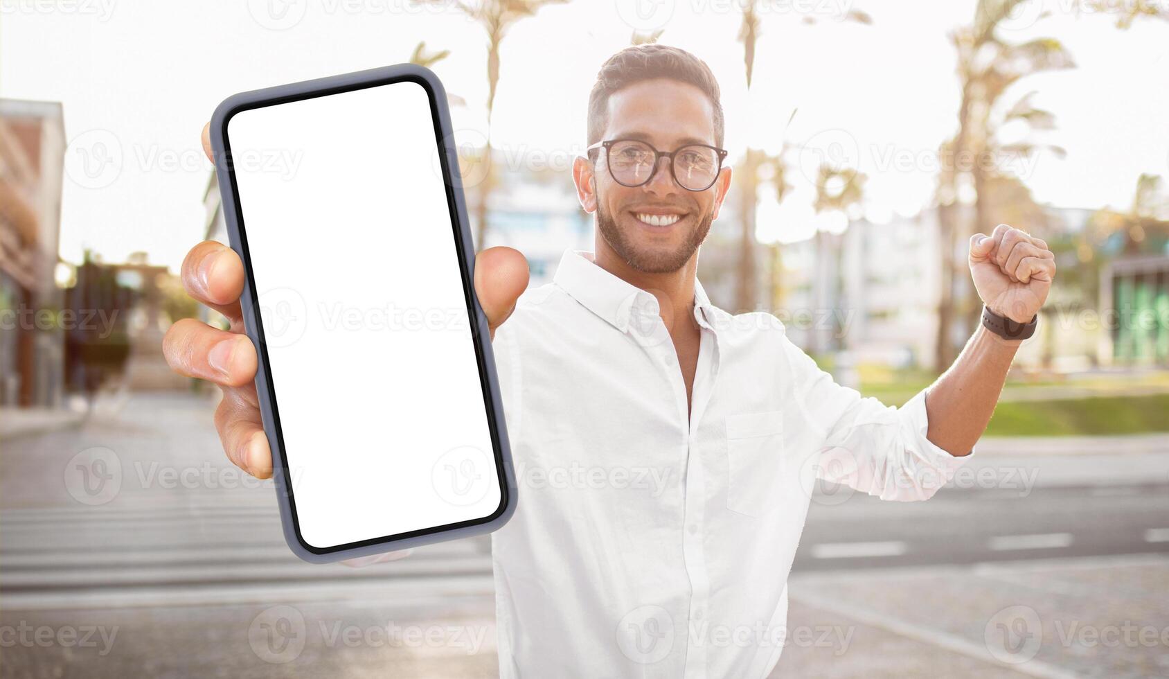 A man in a white shirt is holding a smartphone with a blank screen, celebrating his success. The man is smiling and has his fist raised in the air. He is standing in a city setting photo