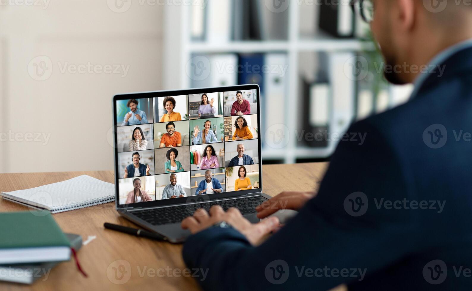 A businessman sits at a desk with a laptop open in front of him, showing a conference screen with many people in individual squares. He is using his laptop for an online meeting. photo