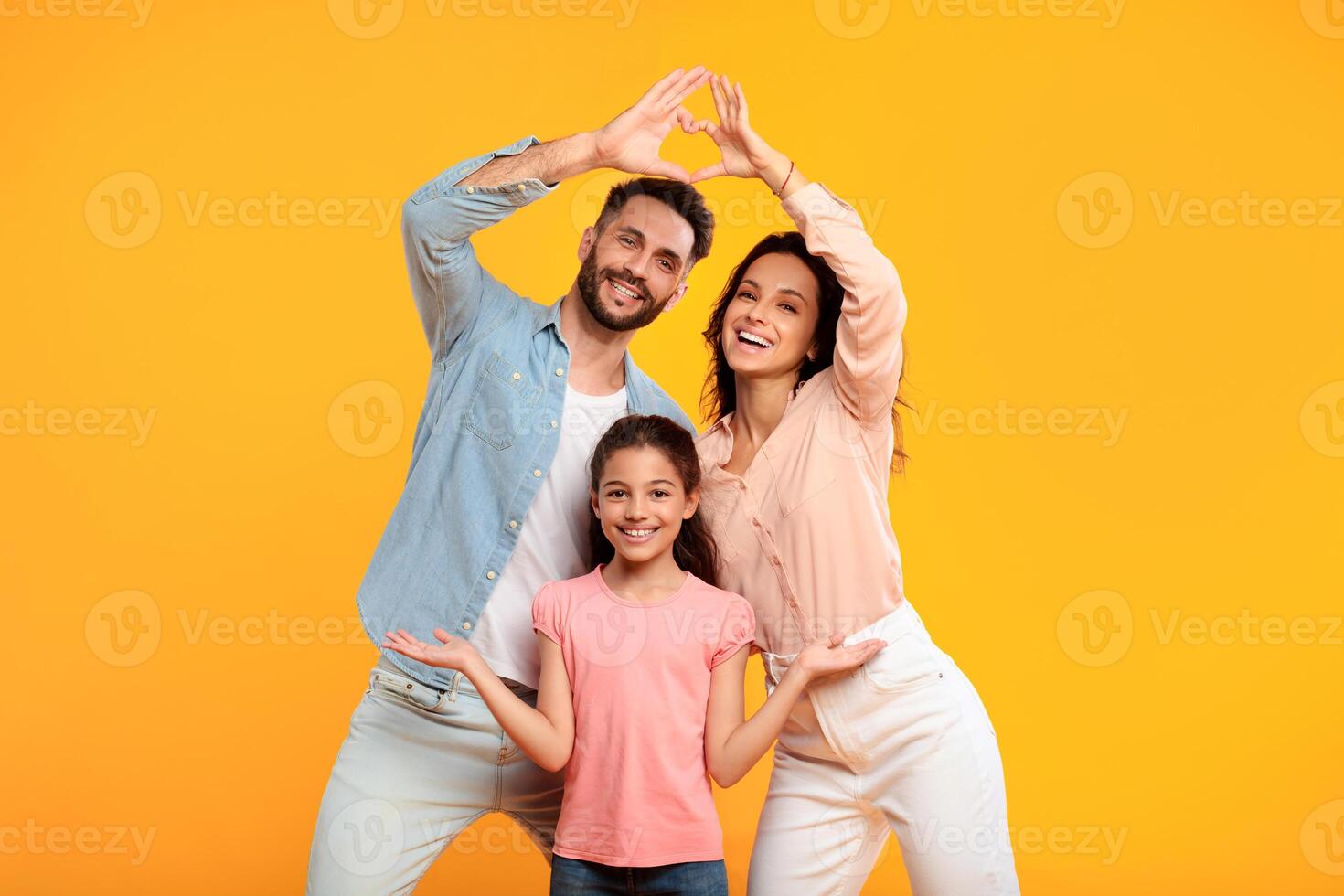 A family of three, a man, woman, and daughter, are standing in front of a bright yellow background. The parents are smiling and making a heart shape with their hands above their daughter's head photo