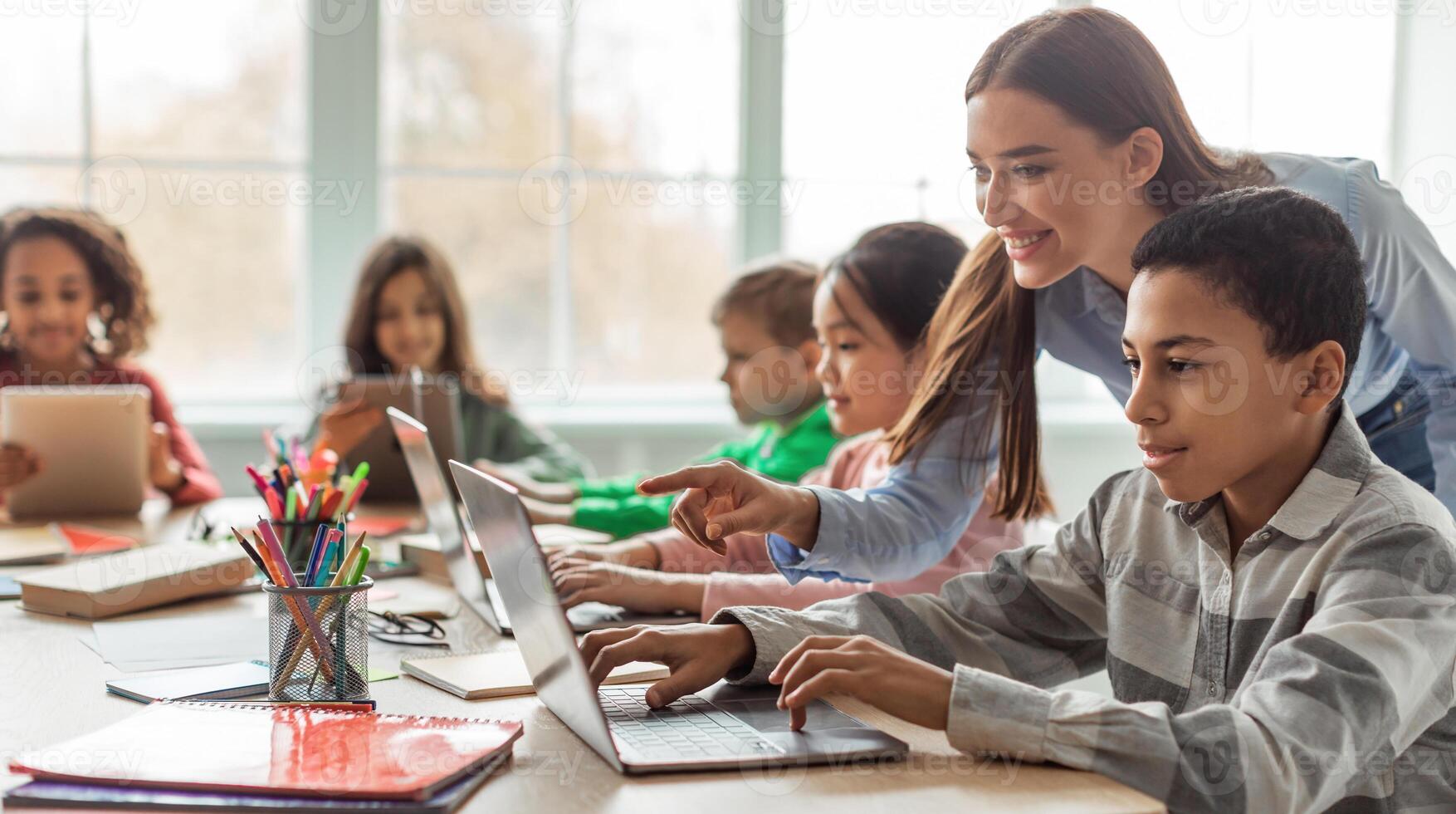 Teacher Teaching Diverse School Kids Using Laptop In Classroom. African American Schoolboy And Diverse Classmates Browsing Internet On Computer Learning Online Indoor. E-Learning Concept photo