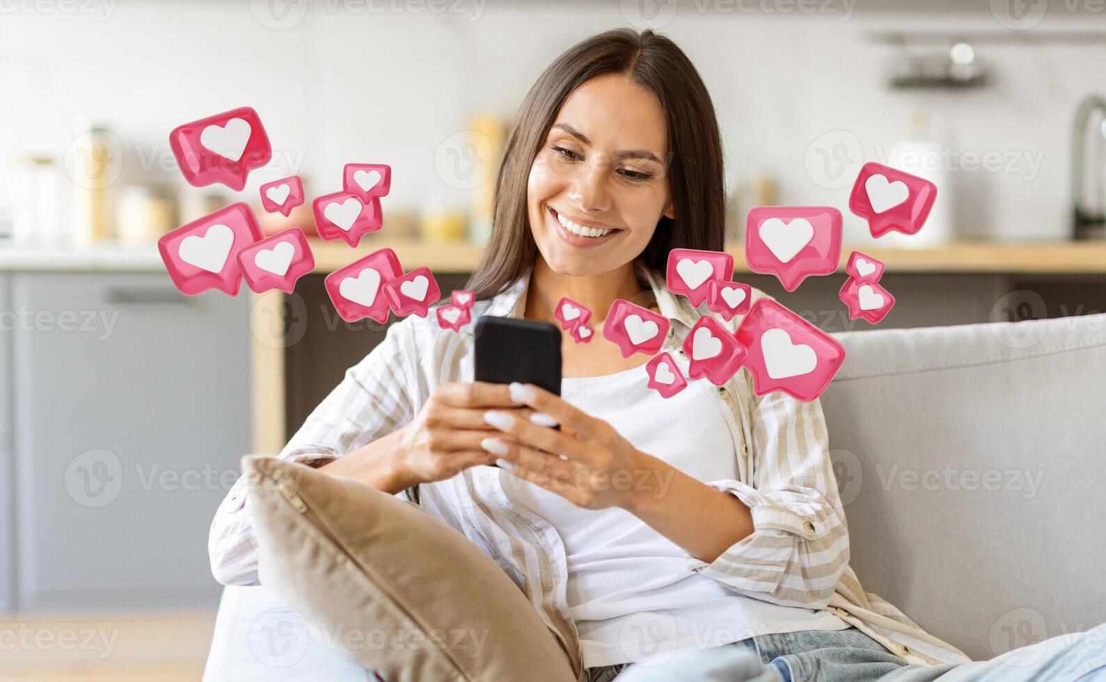 A woman with long brown hair is sitting on a couch in her home. She is smiling as she looks at her smartphone, which is displaying a blank screen. Around her, a number of pink heart icons photo