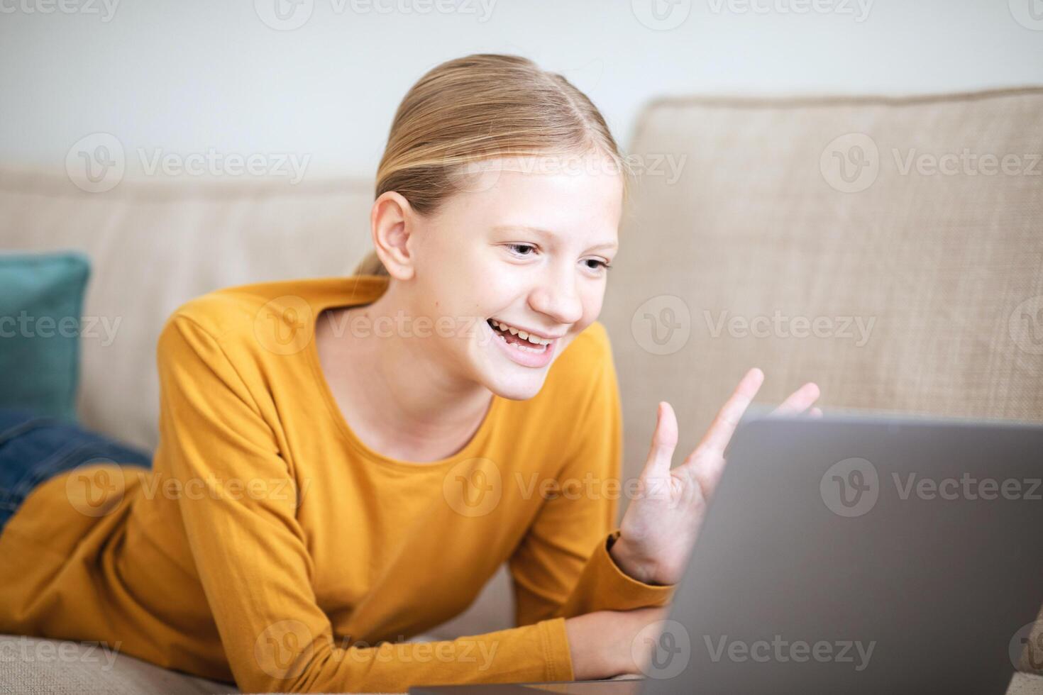 A teen girl in a yellow shirt is lying on a couch and chatting on her laptop. She is smiling and talking, and her hand is raised in a gesture of excitement or emphasis. photo