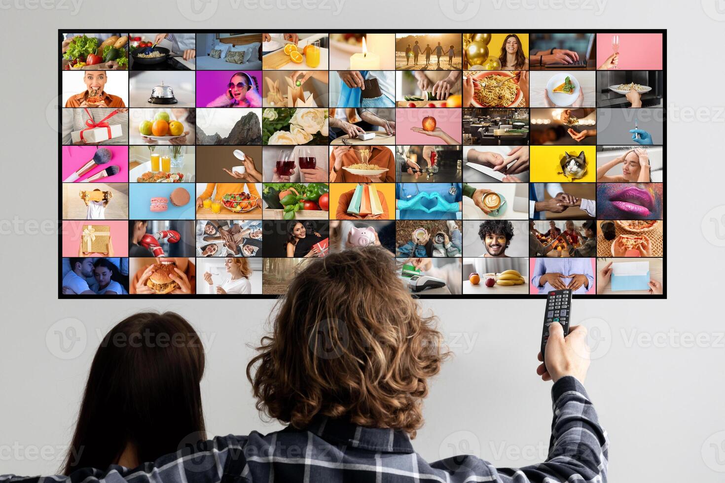 A couple sits on a couch, facing a television screen filled with a collage of images. The woman sits with her back to the camera while the man holds a remote control and faces the screen photo
