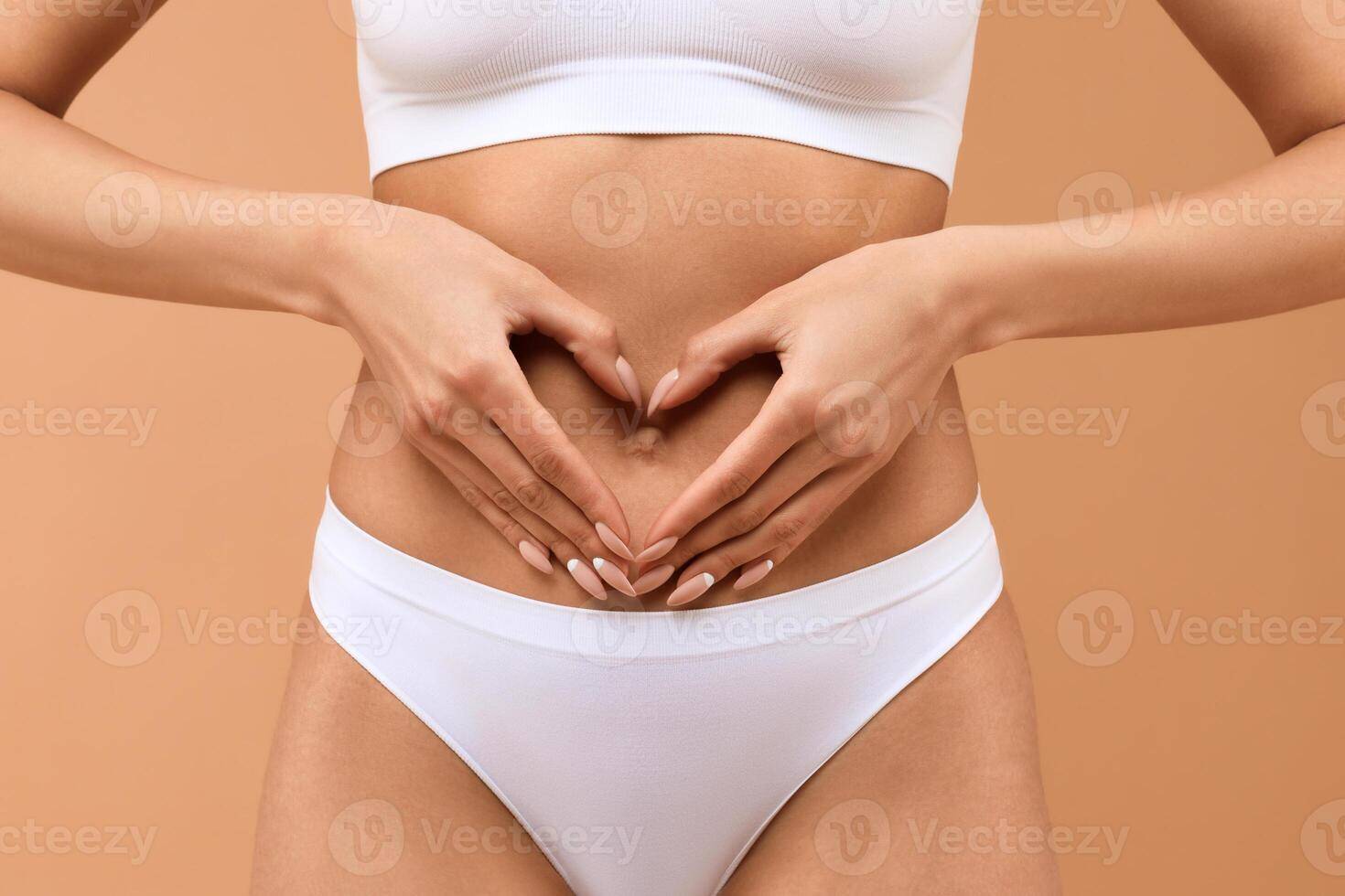 Cropped of woman wearing white underwear holding hands in heart shaped gesture over her belly, beige studio background photo