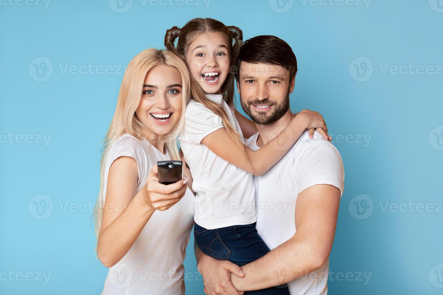 A happy family is watching television together at home. The parents are smiling and the daughter is holding onto her father while watching the screen. photo