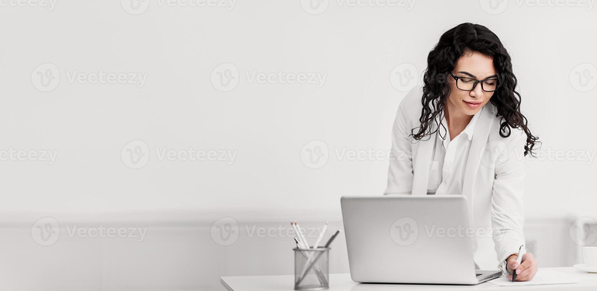 Note Taking. Happy mexican businesswoman in glasses at work, standing at desk and writing to do list, copyspace photo