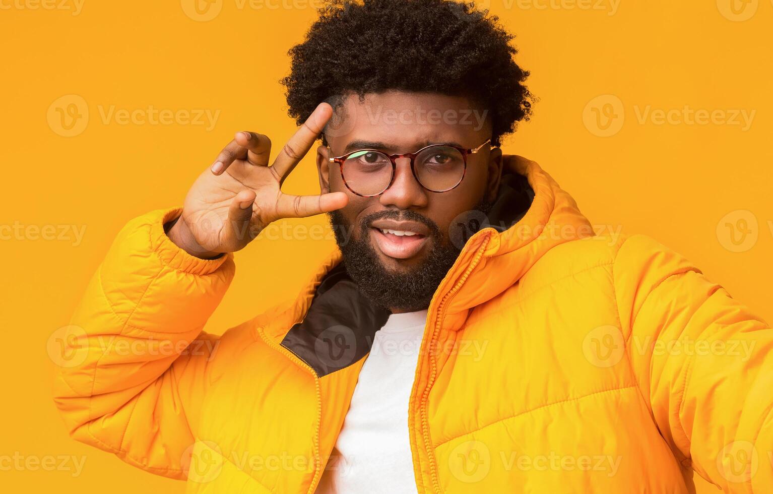 Handsome winter afro guy making selfie over orange background, wearing glasses and showing peace symbol next to eye photo