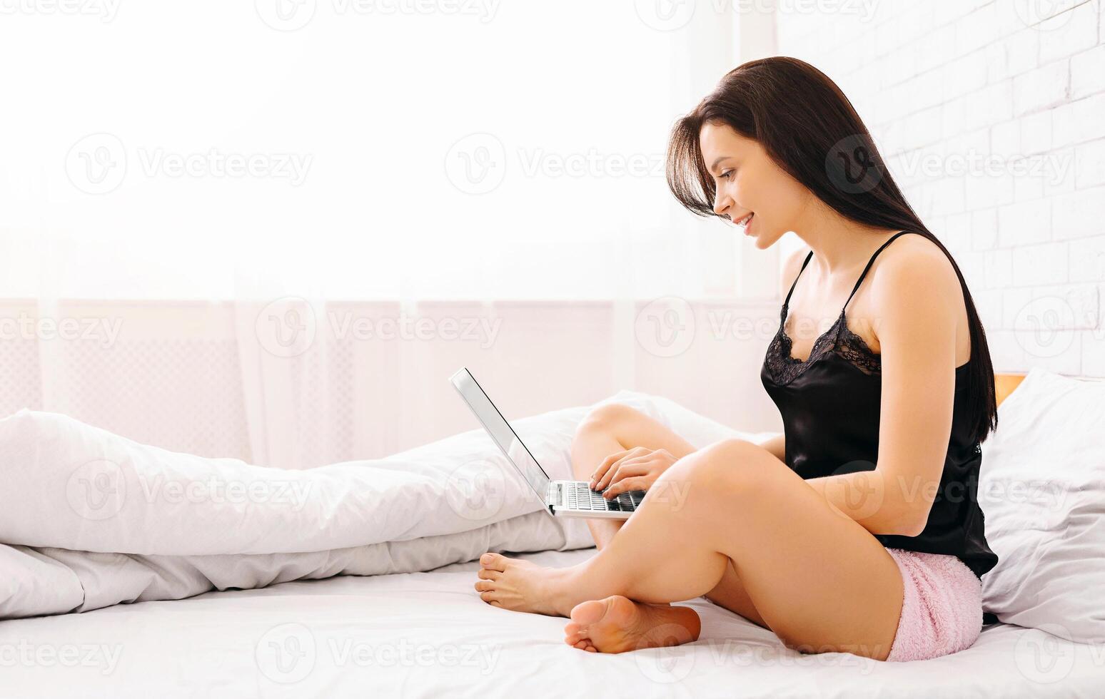 A woman is seated on a bed, engaging with a laptop computer. She appears focused and concentrated on the screen as she types or reads. The room is softly lit, creating a cozy atmosphere. photo