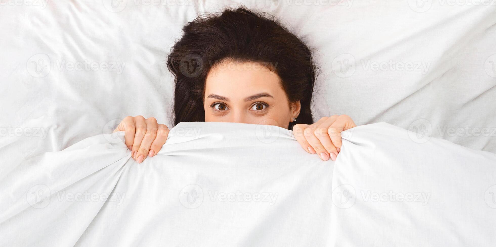 A woman is crouched underneath a blanket on a bed, trying to shield herself from view. Her body is partially hidden, with only her head peeking out from the covers. photo