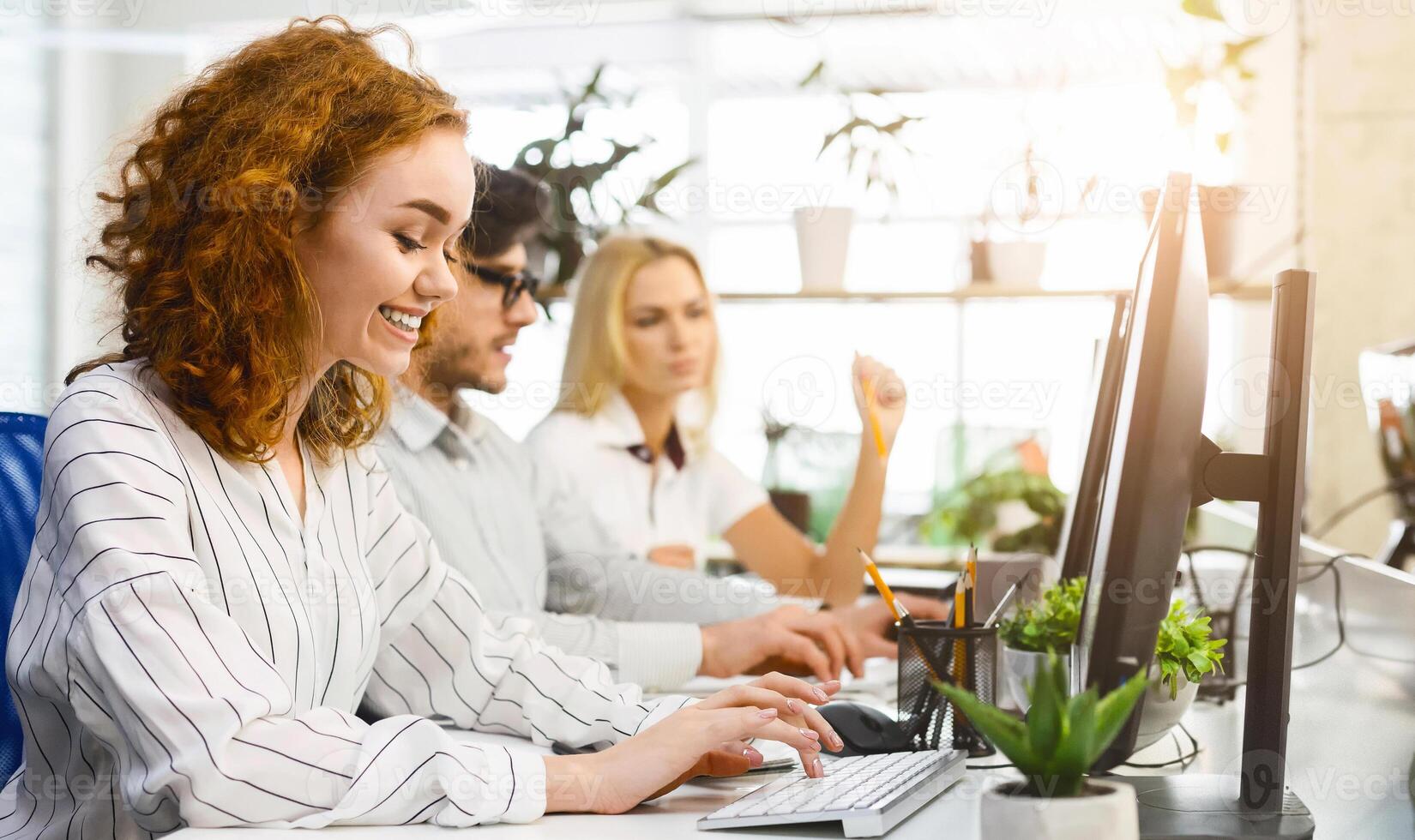 Several coworkers are seated at a desk, each engrossed in their computer screens, actively typing and clicking. The group appears focused and concentrated on their tasks photo