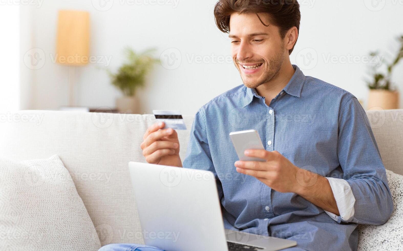 A man sits on a couch, smiling as he makes a payment online using a credit card and smartphone. photo