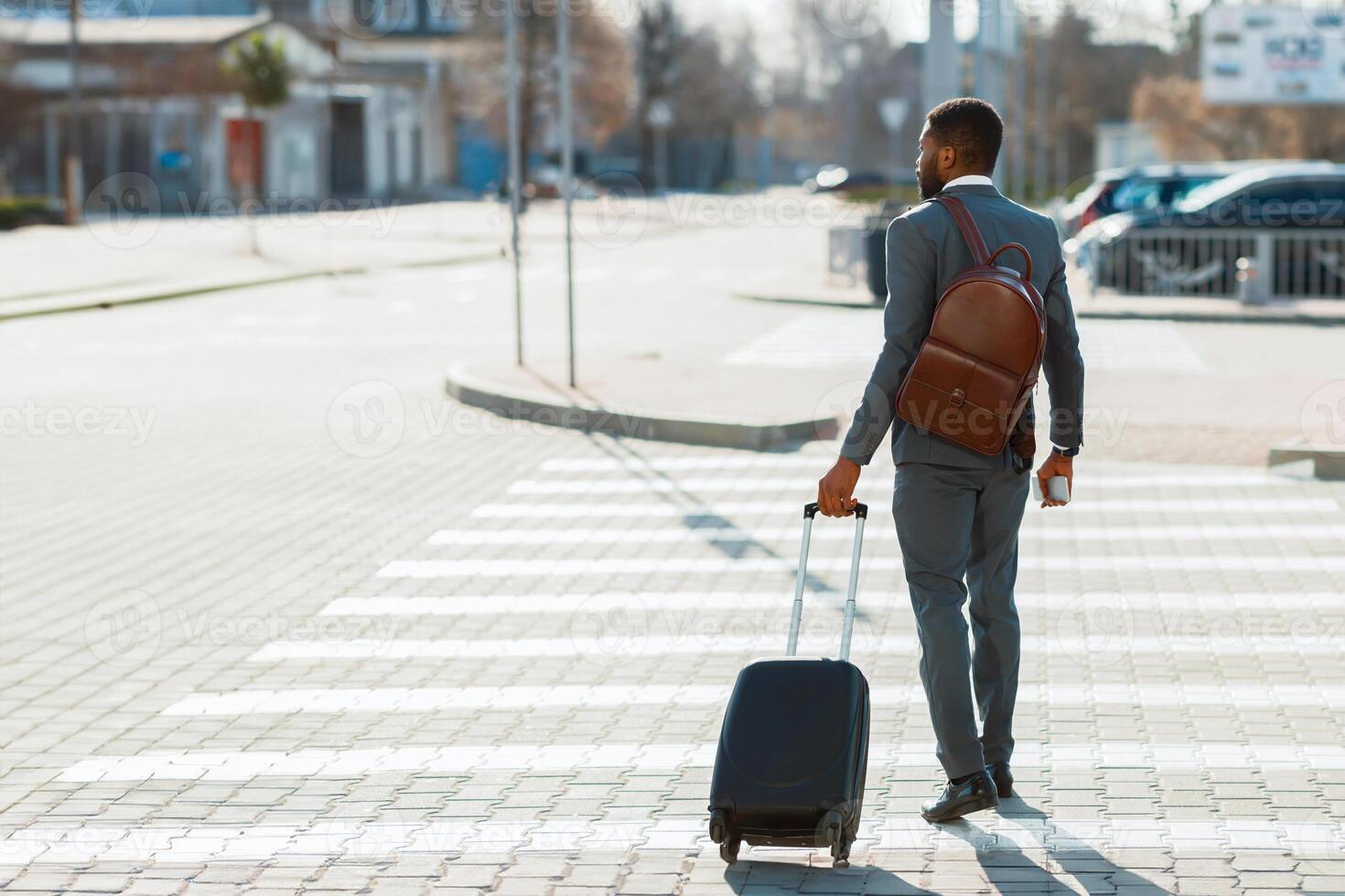 African American businessman in a suit crossing the street with a rolling suitcase, seen from behind in an urban setting, back view photo
