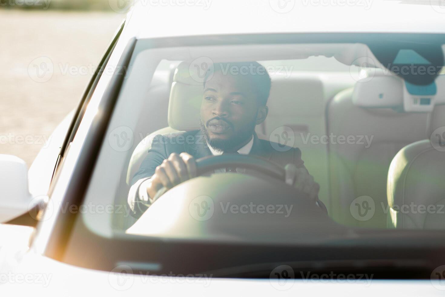 Black man wearing a suit sits in the drivers seat of a car. The car is facing towards the sun, creating a bright, hazy effect. He is gripping the steering wheel and looking straight ahead photo