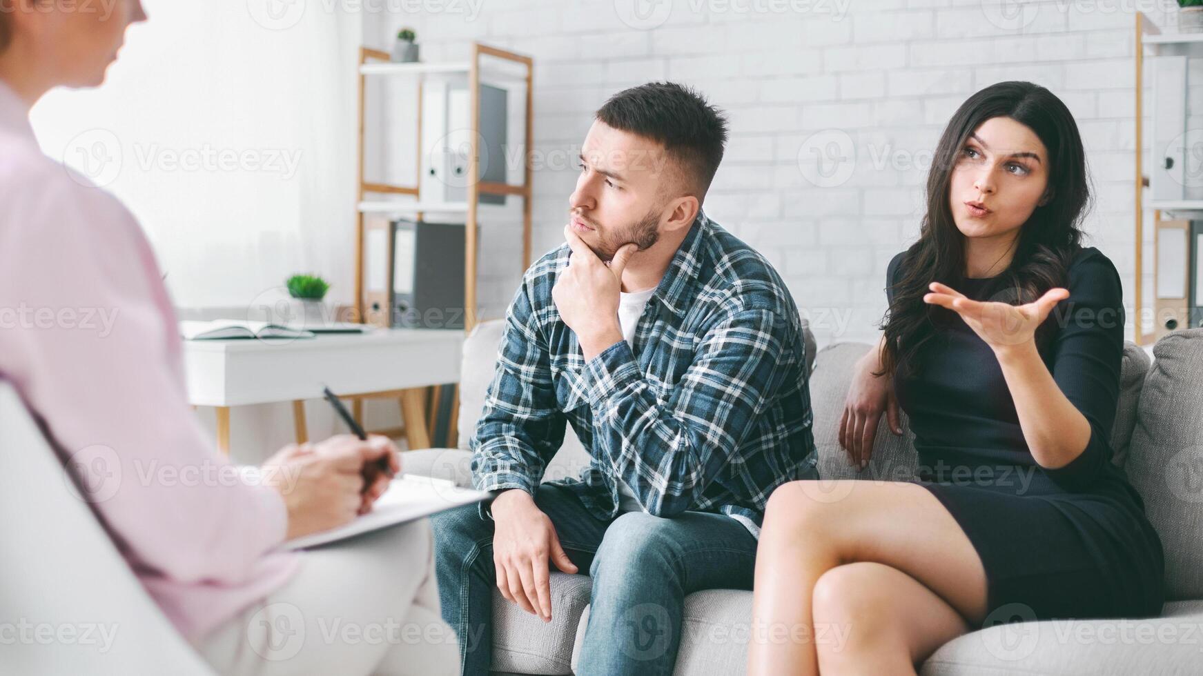 A couple sits on a couch in an office, talking to a therapist. The woman is talking and gesturing with her hand, while the man is listening and thinking thoughtfully. The therapist is taking notes. photo
