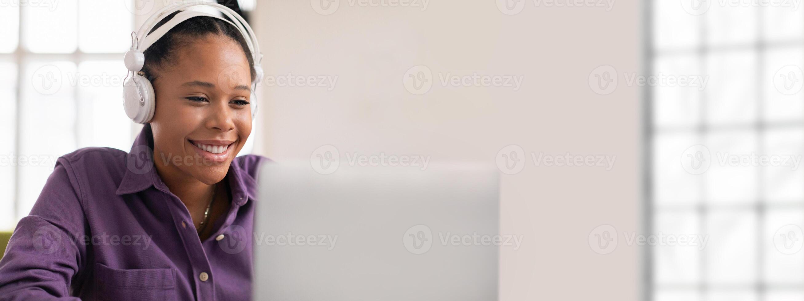 A young African American woman wearing headphones smiles while participating in a virtual meeting. The bright office setting features large windows that flood the room with natural light. photo