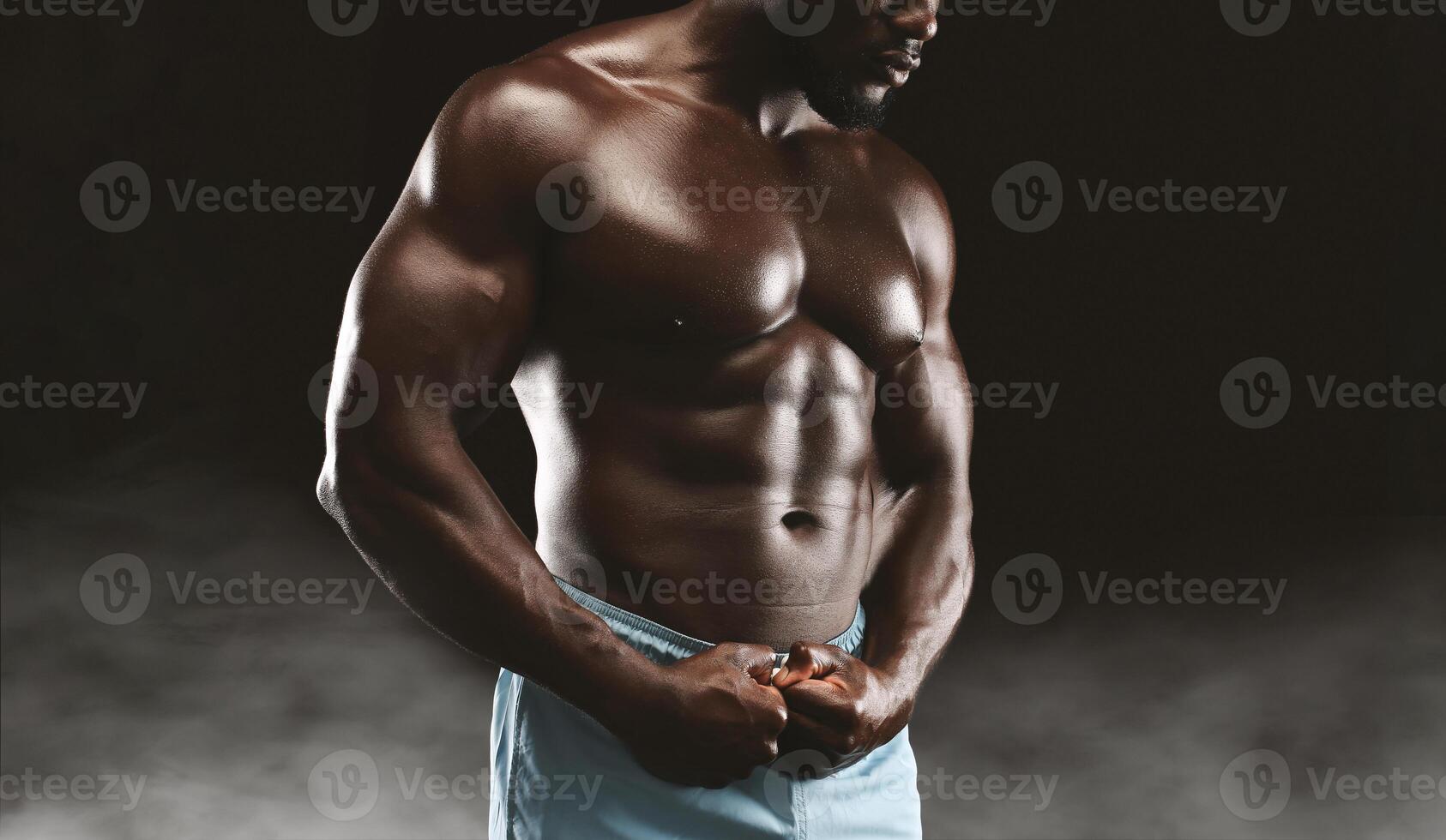 A muscular man posing in studio, displaying defined abs and biceps in shorts against dark background for dramatic effect. photo
