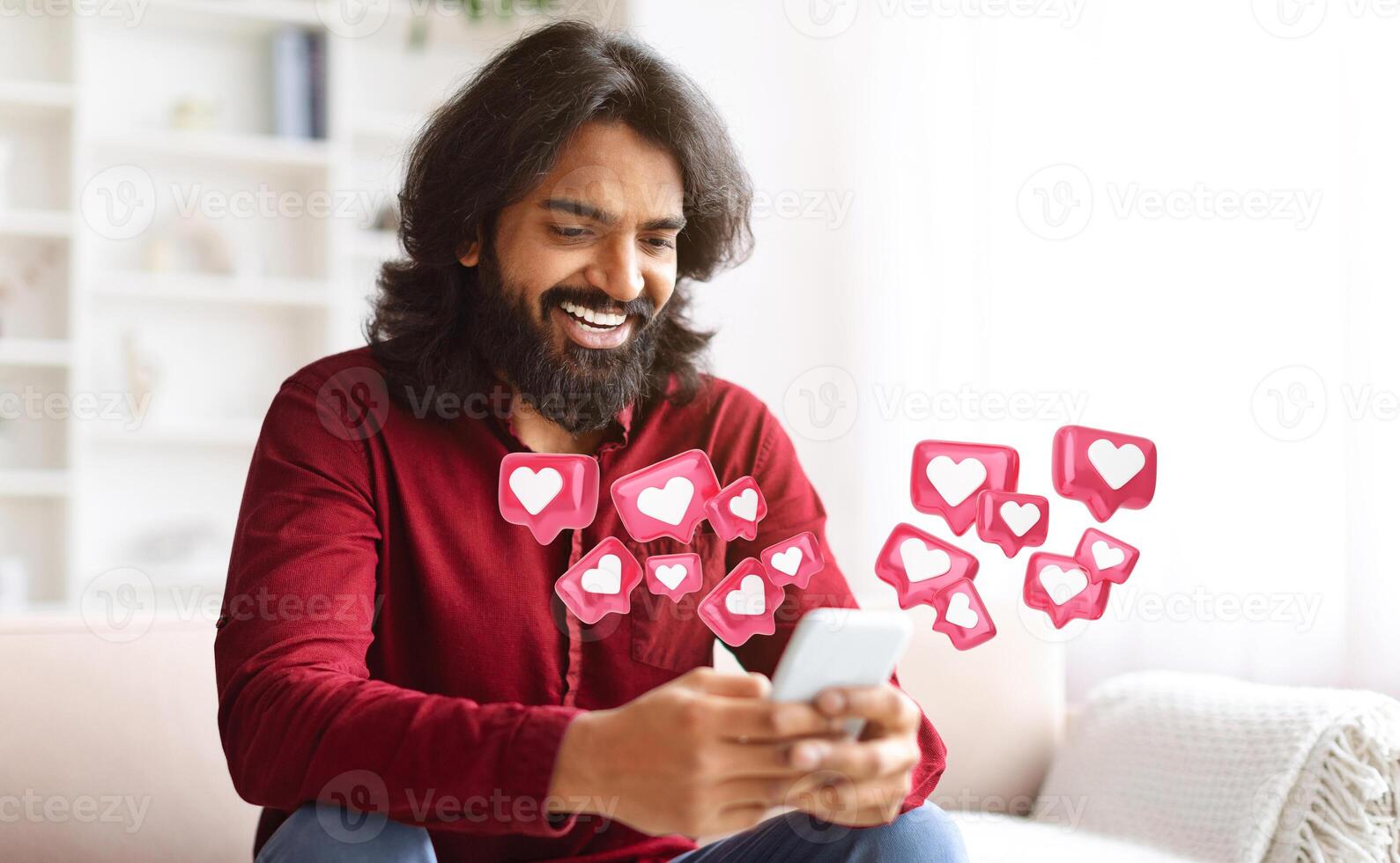 Indian man with a beard and long hair sits on a couch, smiling while looking at his smartphone. Surrounding him are digital hearts, symbolizing likes or social media engagement photo