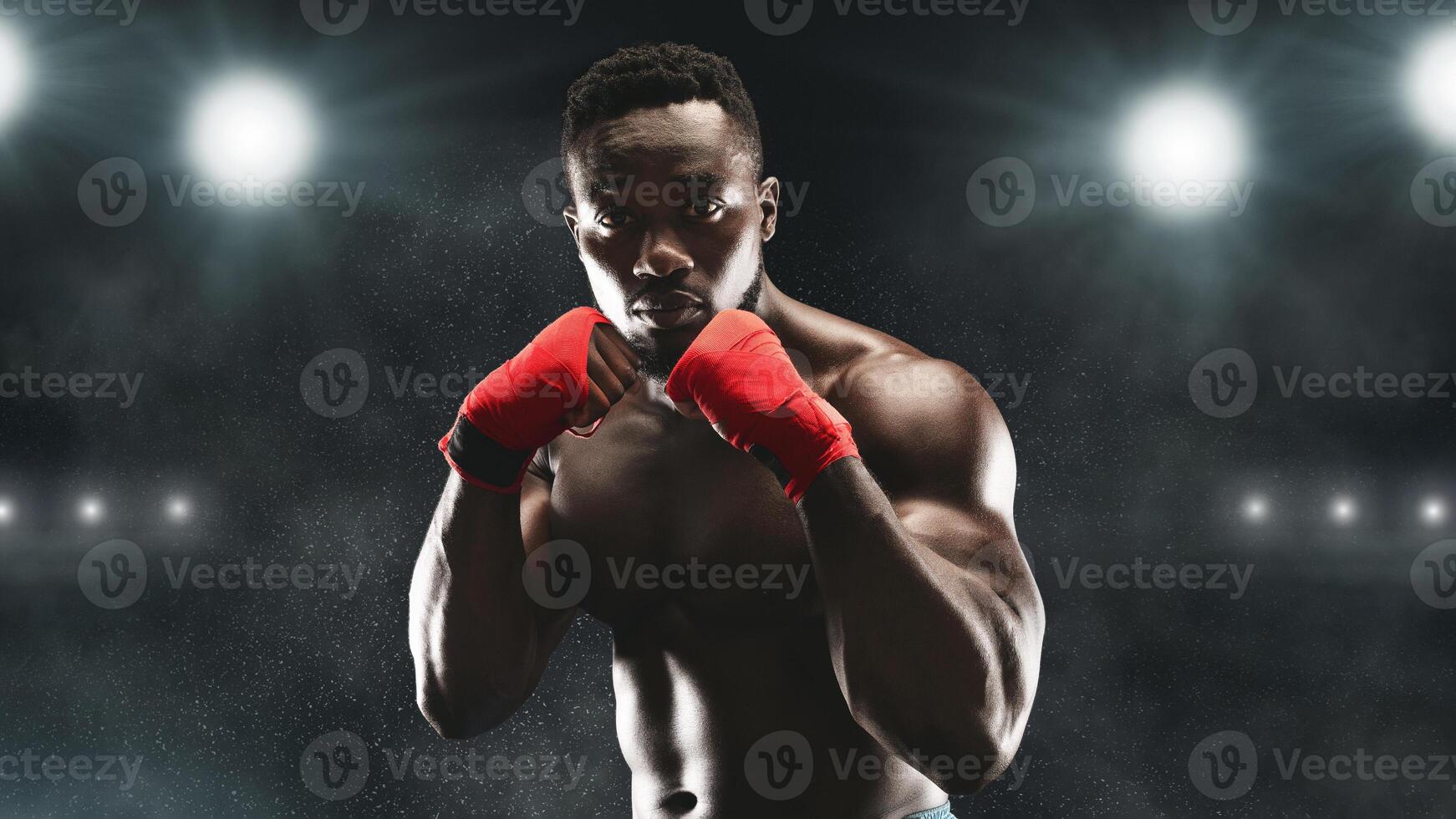 A boxer in red hand wraps stands ready in a dark stadium, illuminated by bright lights. photo