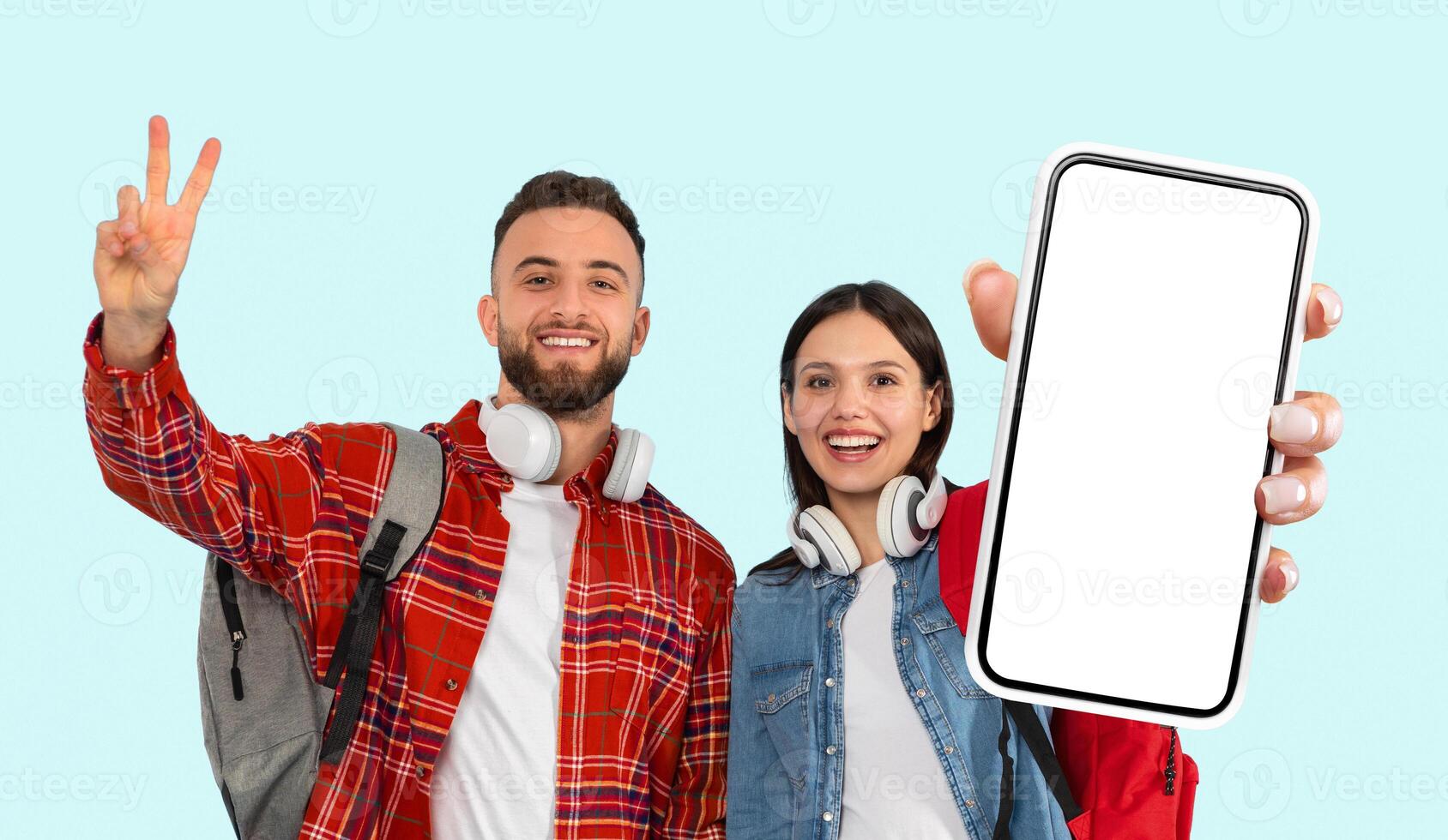 A young man and woman stand in front of a light blue background. The man is wearing a red plaid shirt and a backpack and has headphones around his neck. photo