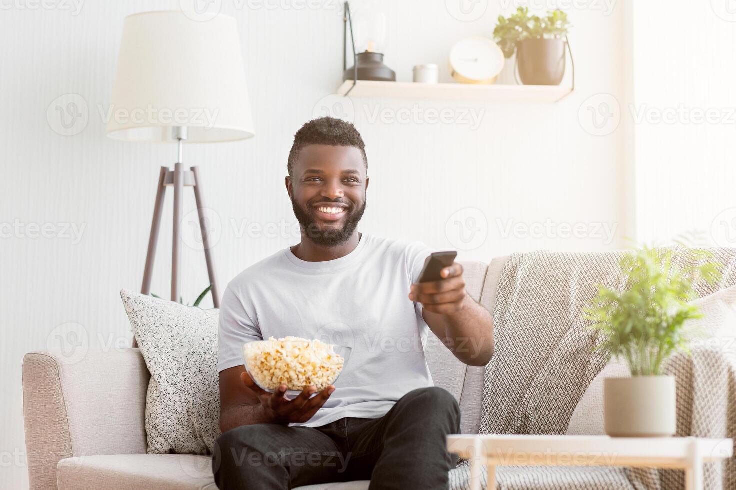 Black man is sitting on a couch in a living room. He is holding a bowl of popcorn and a remote control in his hands. He is smiling and looking at the television photo