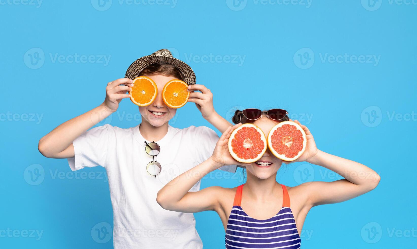 Two children, a boy wearing a hat and a girl in a striped swimsuit, playfully hold orange and grapefruit slices over their eyes against a blue background. photo