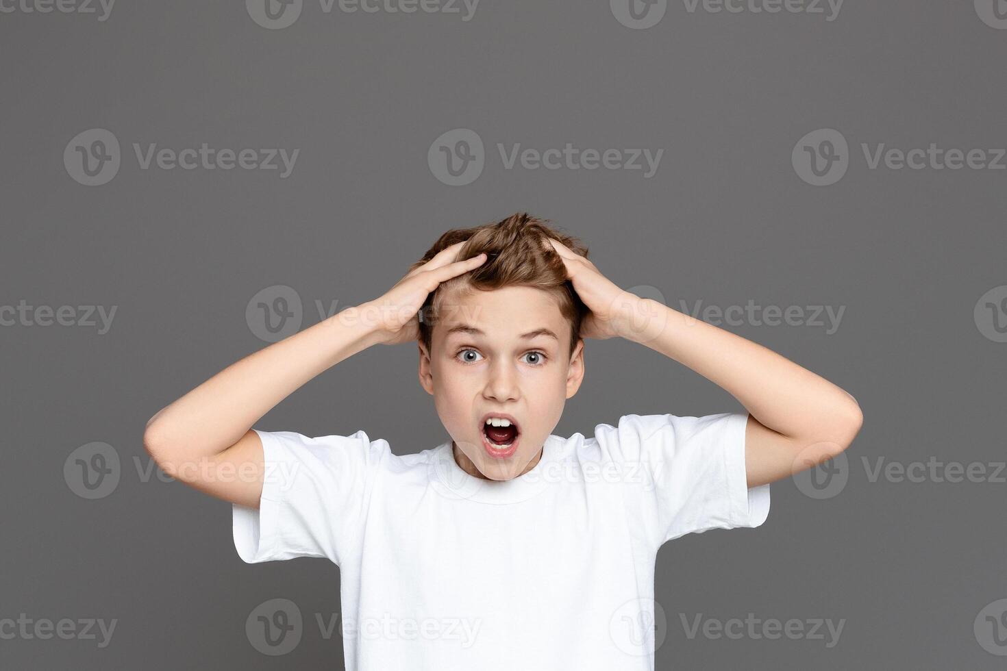 A shocked teen boy is captured with wide eyes and hands clutching his head against an isolated grey backdrop, suggesting alarm photo