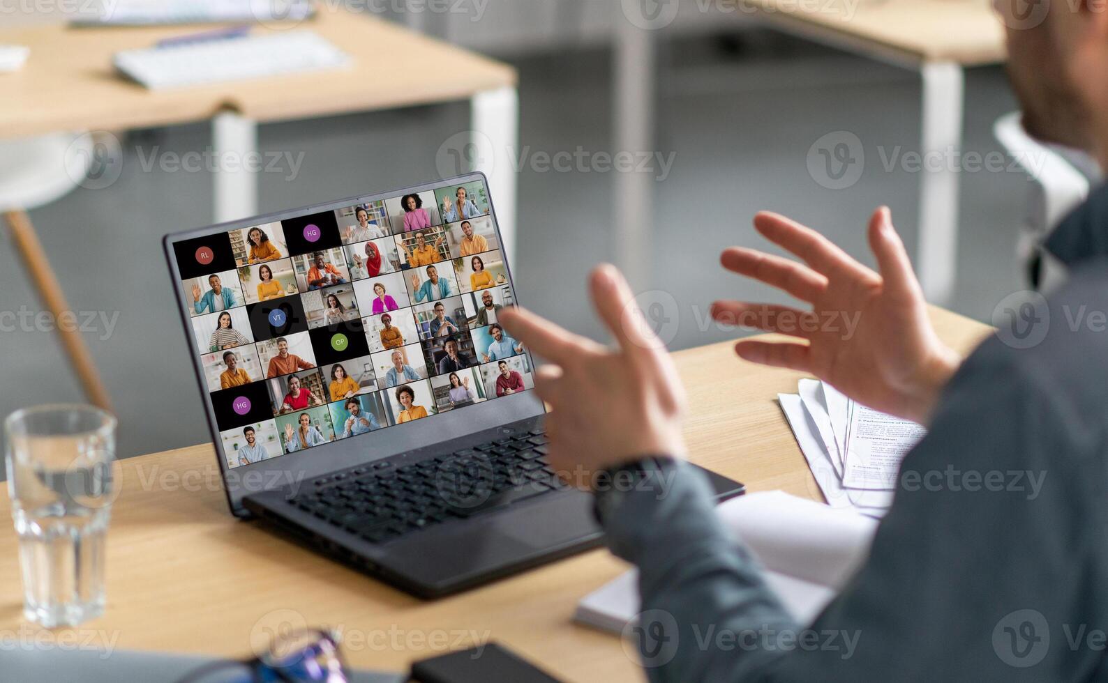 A man sits at a desk in front of a laptop. He is gesturing with his hands and looking at the screen, which displays a grid of calls with multiple people. photo