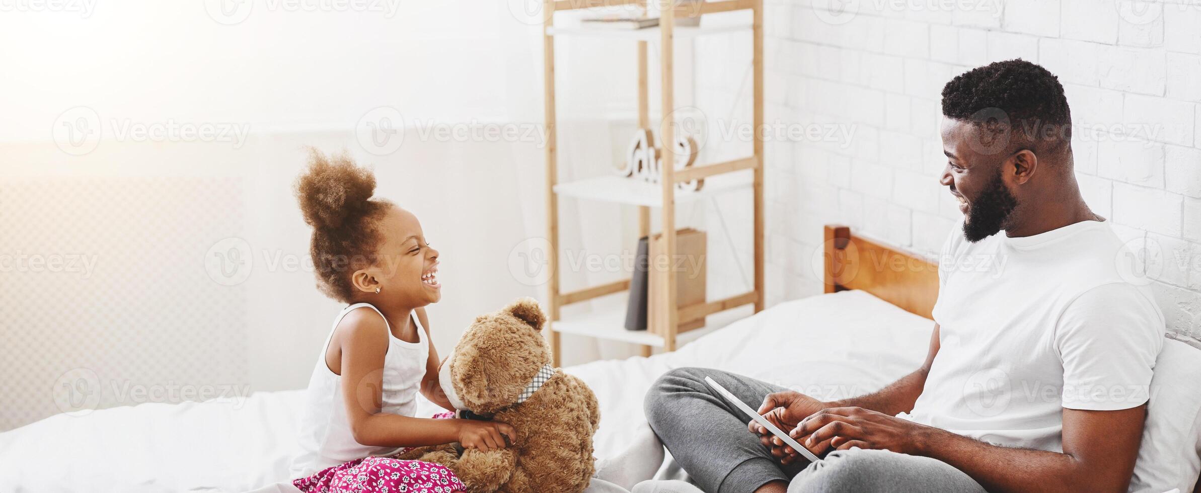 A little black girl is sitting on a bed, hugging a teddy bear and smiling at her dad. Her dad is sitting next to her, using a tablet and smiling back at her. photo