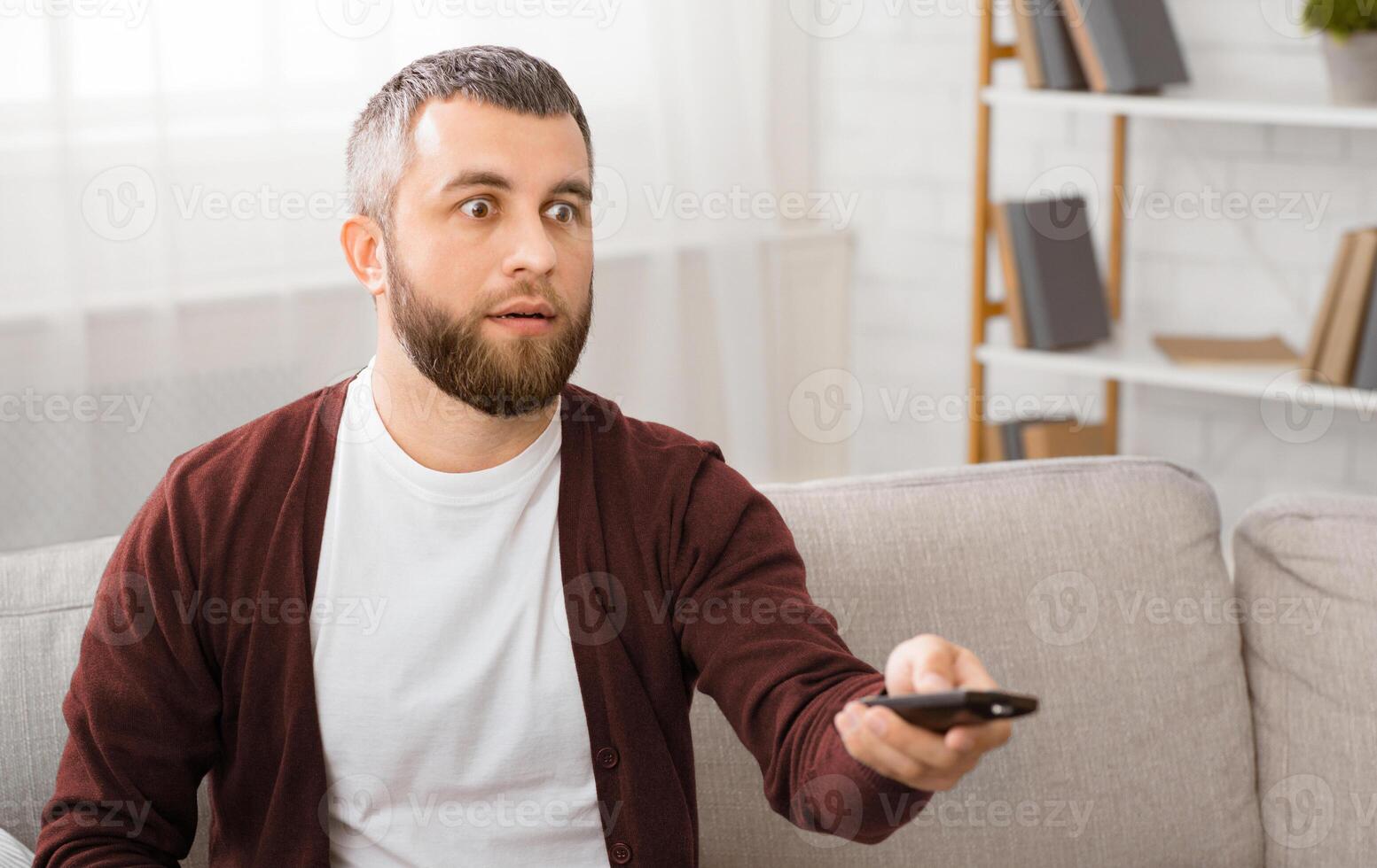 A man is seated on a couch, with a remote control in his hand. He appears relaxed and focused on the device, presumably changing channels or adjusting volume on the television. photo