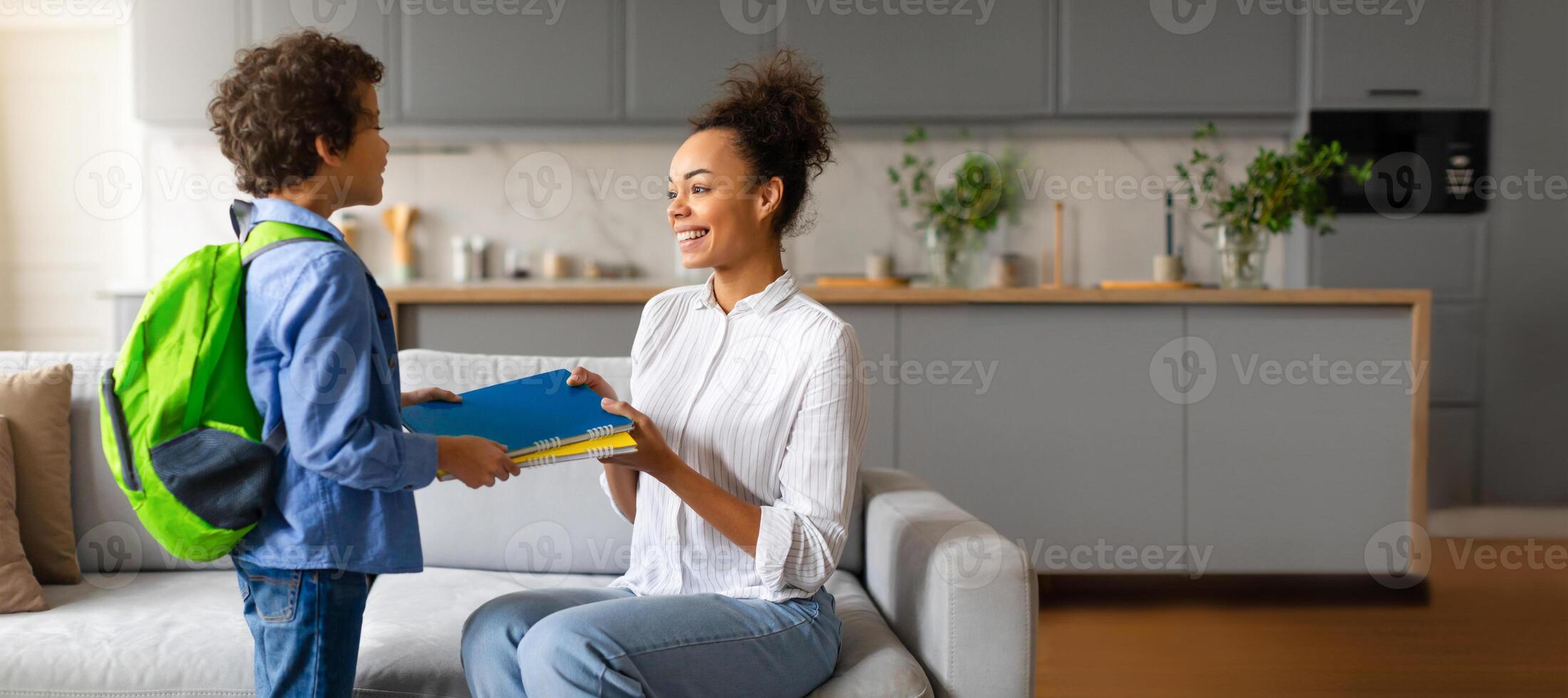 African American mother smiles as she hands a book to her young son, who holds a backpack, in a warm and inviting kitchen. Sunlight brightens the space, creating a joyful atmosphere for learning. photo