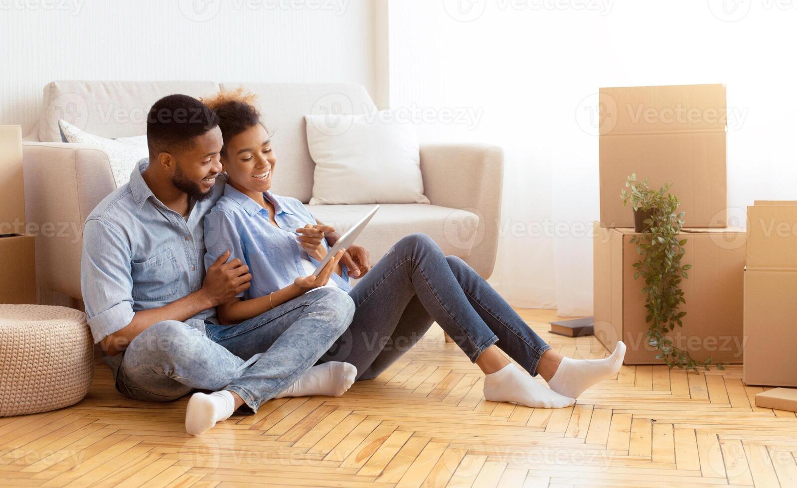 Moving Apartment. Cheerful African American Spouses Using Tablet Computer Sitting On Floor Among Unpacked Boxes In New Flat. photo