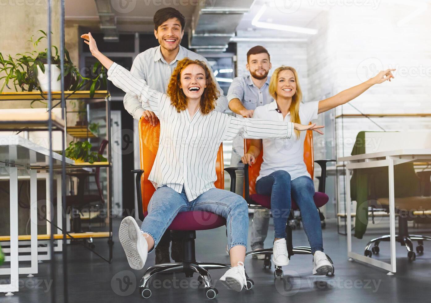 Time for break. Happy colleagues having fun at workplace, making office chair race in coworking space photo