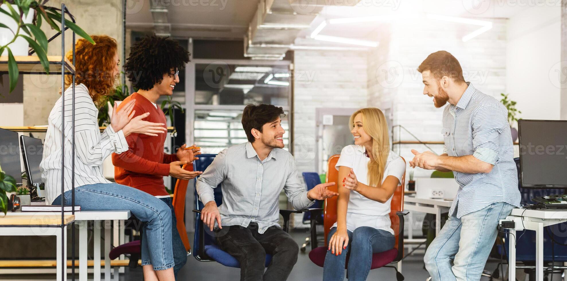 A group of young professionals is gathered in a brightly lit office space, engaging in an animated discussion. Some members are seated while others stand, indicating collaborative work environment photo