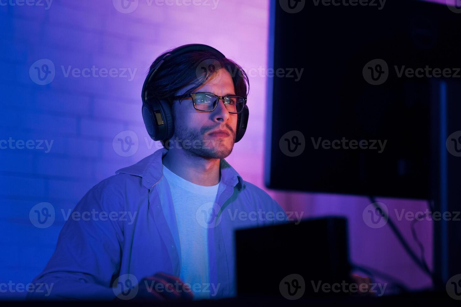 A man with headphones on is intently focused on his computer screen in a room illuminated by purple and blue lighting. He is wearing glasses and a light blue button-down shirt. photo