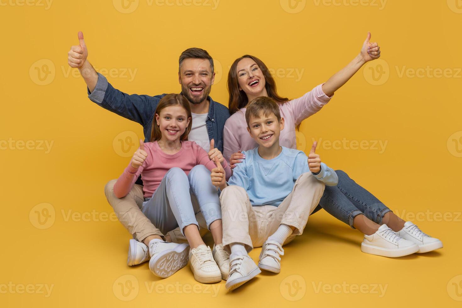 A smiling family consisting of a mother, father, daughter, and son are sitting close to each other against a vibrant yellow backdrop, all giving a thumbs-up sign in a show of happiness and unity. photo