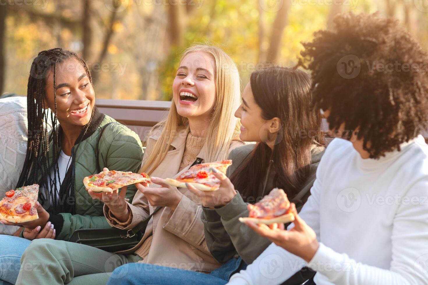Cheerful multiracial teens joking and laughing while eating pizza, sitting on bench in park photo