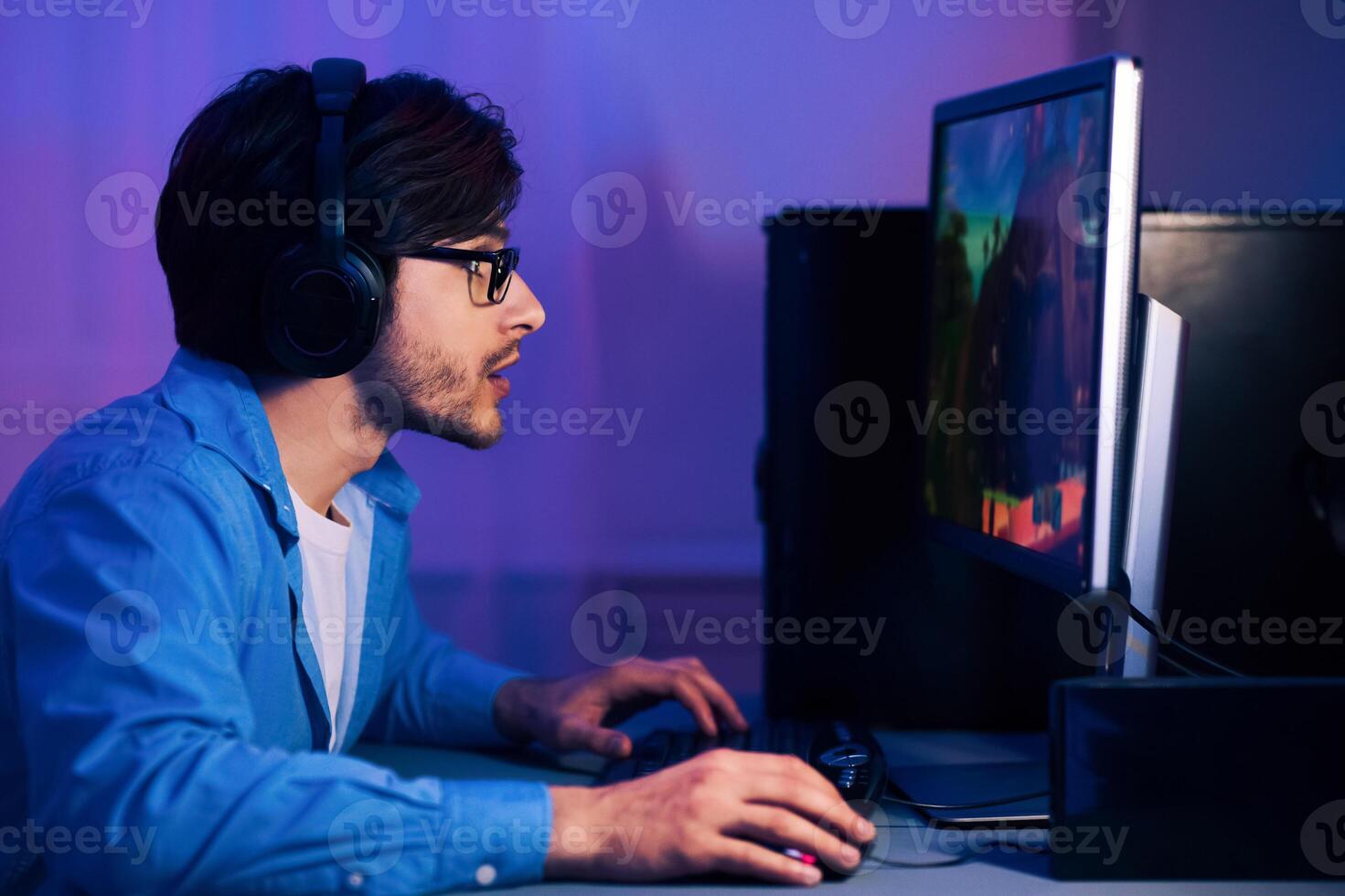 A young man wearing glasses and headphones sits at his computer, focused on playing a game. His room is dimly lit with blue and purple hues, creating an atmospheric ambiance. photo