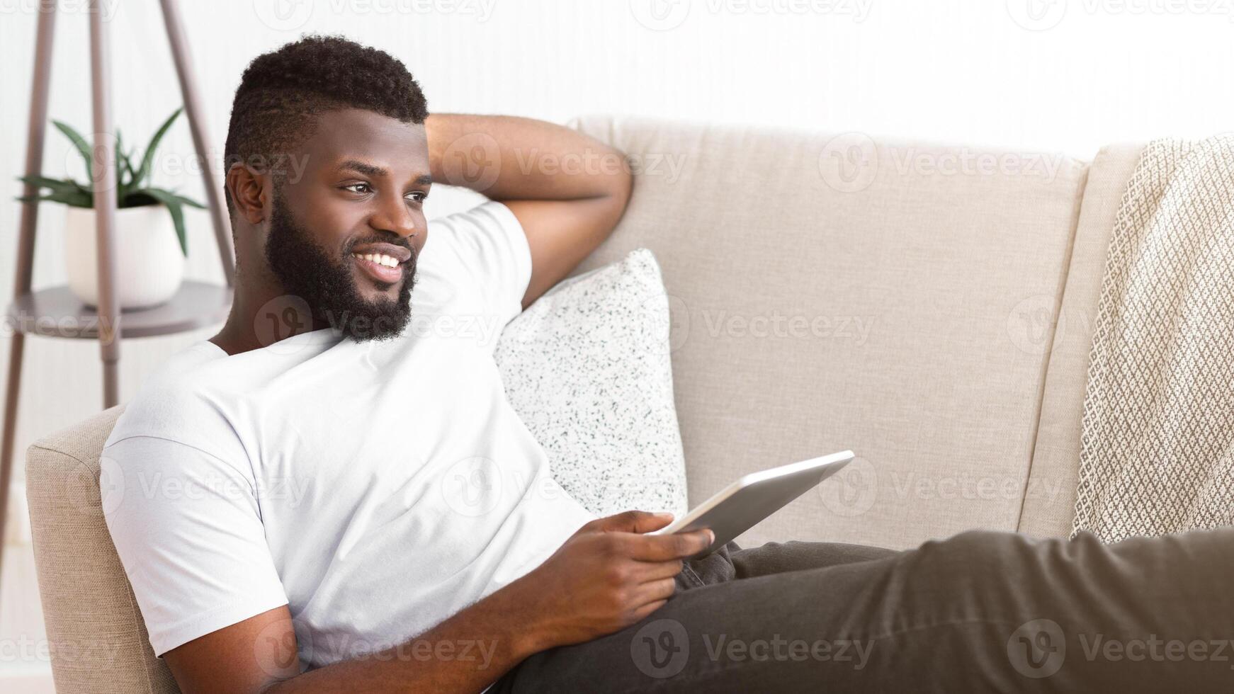 African American man with a beard is sitting on a couch, leaning back, while using a tablet. He is wearing a white shirt and gray pants. He is looking to the side with a smile on his face. photo