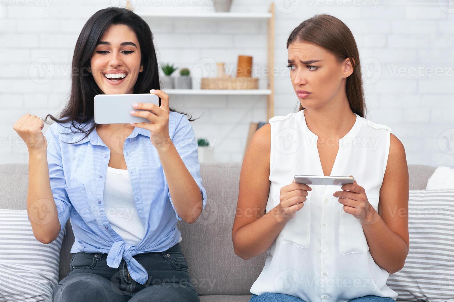 Envy, Competition And Friendship. Girl Being Upset After Losing Mobile Game Sitting On Couch With Friend Indoor. photo