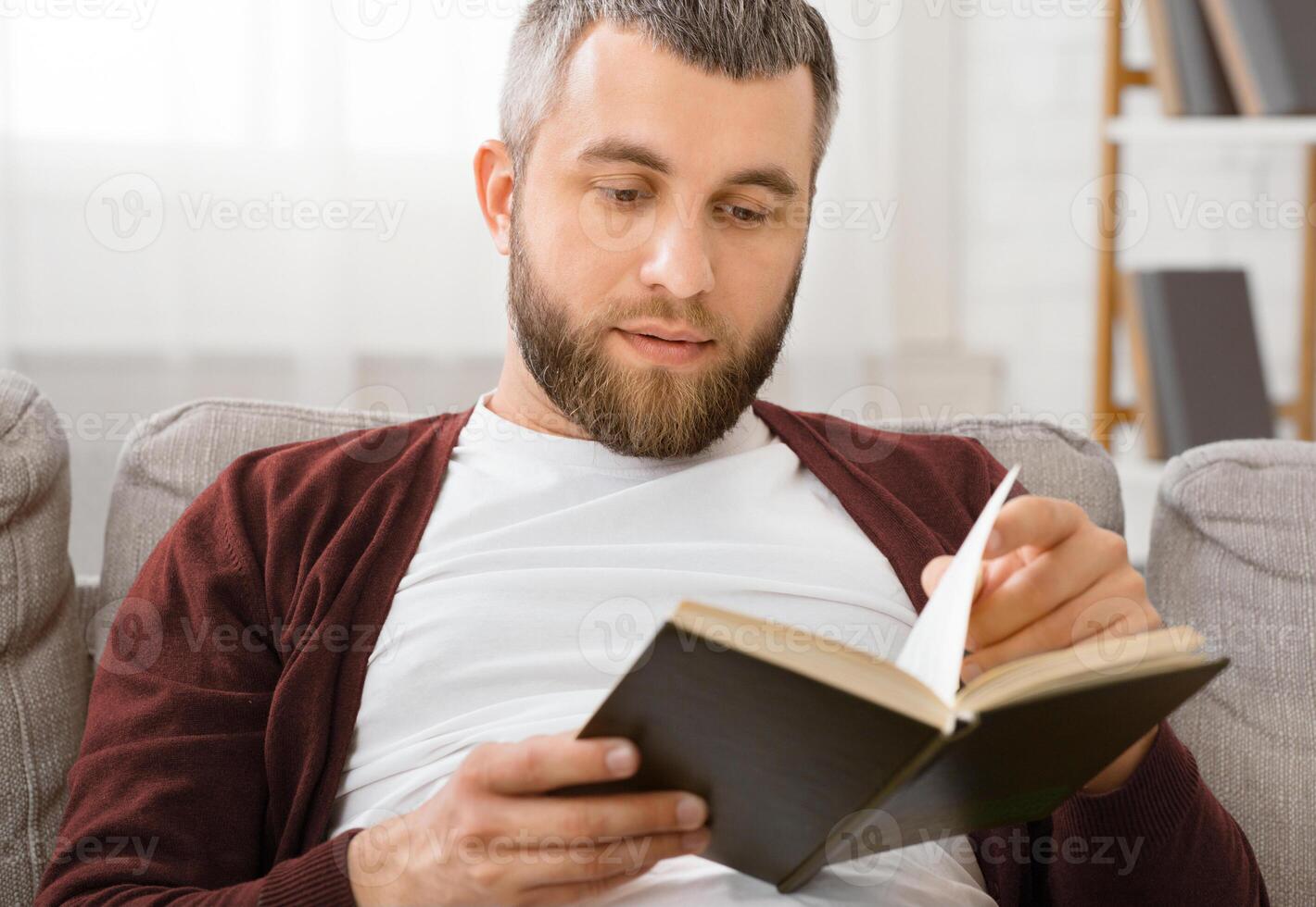 A man is seated on a couch, engrossed in reading a book. He is focused and turning the pages, with a concentrated expression on his face. The room is well-lit, creating a cozy reading environment. photo