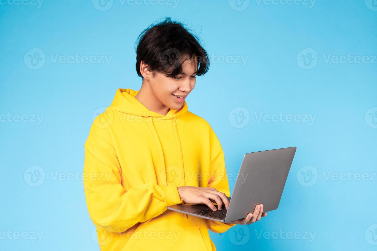 Asian teen guy wearing a yellow hoodie is sitting at a laptop, typing on the keyboard. The laptop screen displays a webpage or document. The person appears focused on their work. photo
