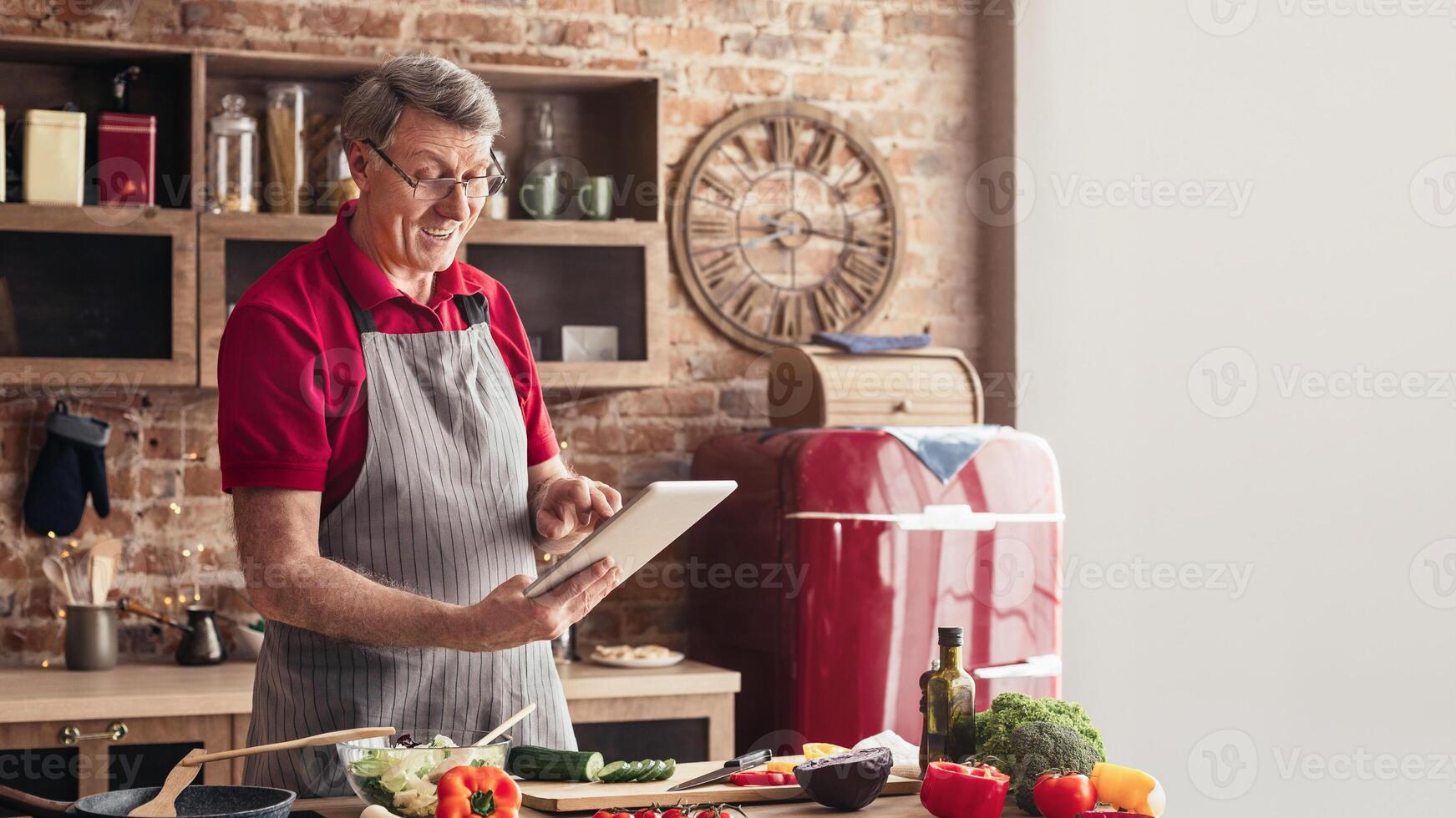 A senior man in a red shirt and apron stands in his kitchen, holding a tablet and smiling. He is following a recipe while surrounded by fresh vegetables, a cutting board, and a red refrigerator photo