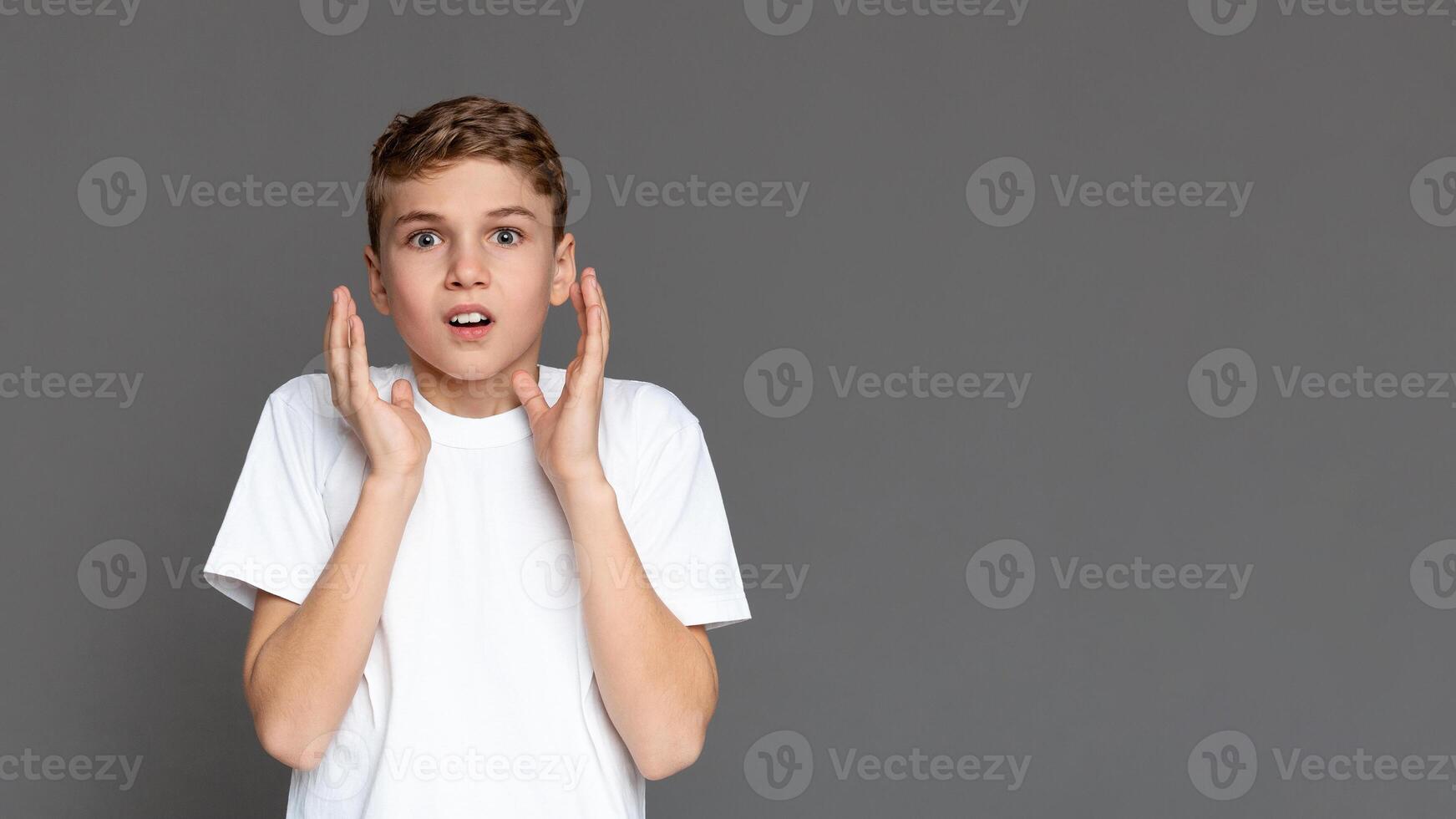 A teenager with a shocked expression holds his cheeks isolated against a gray background, showing a range of youngster's emotions, copy space photo