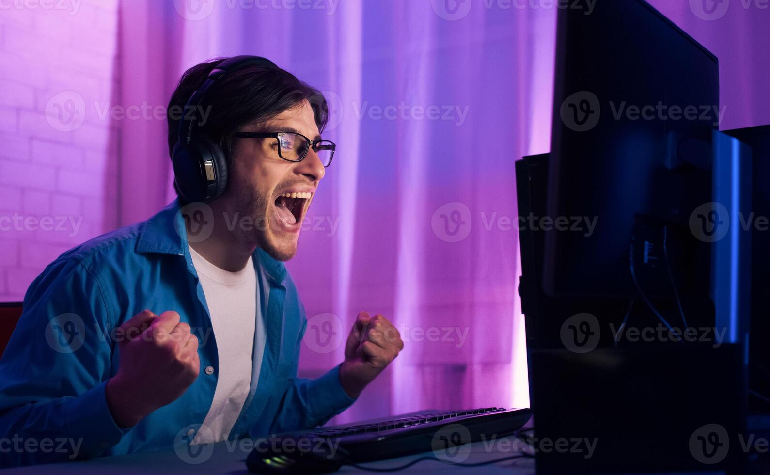 A young man wearing headphones and glasses is seated in front of a computer screen playing games. He is excited and has his arms raised in the air as if in victory. photo